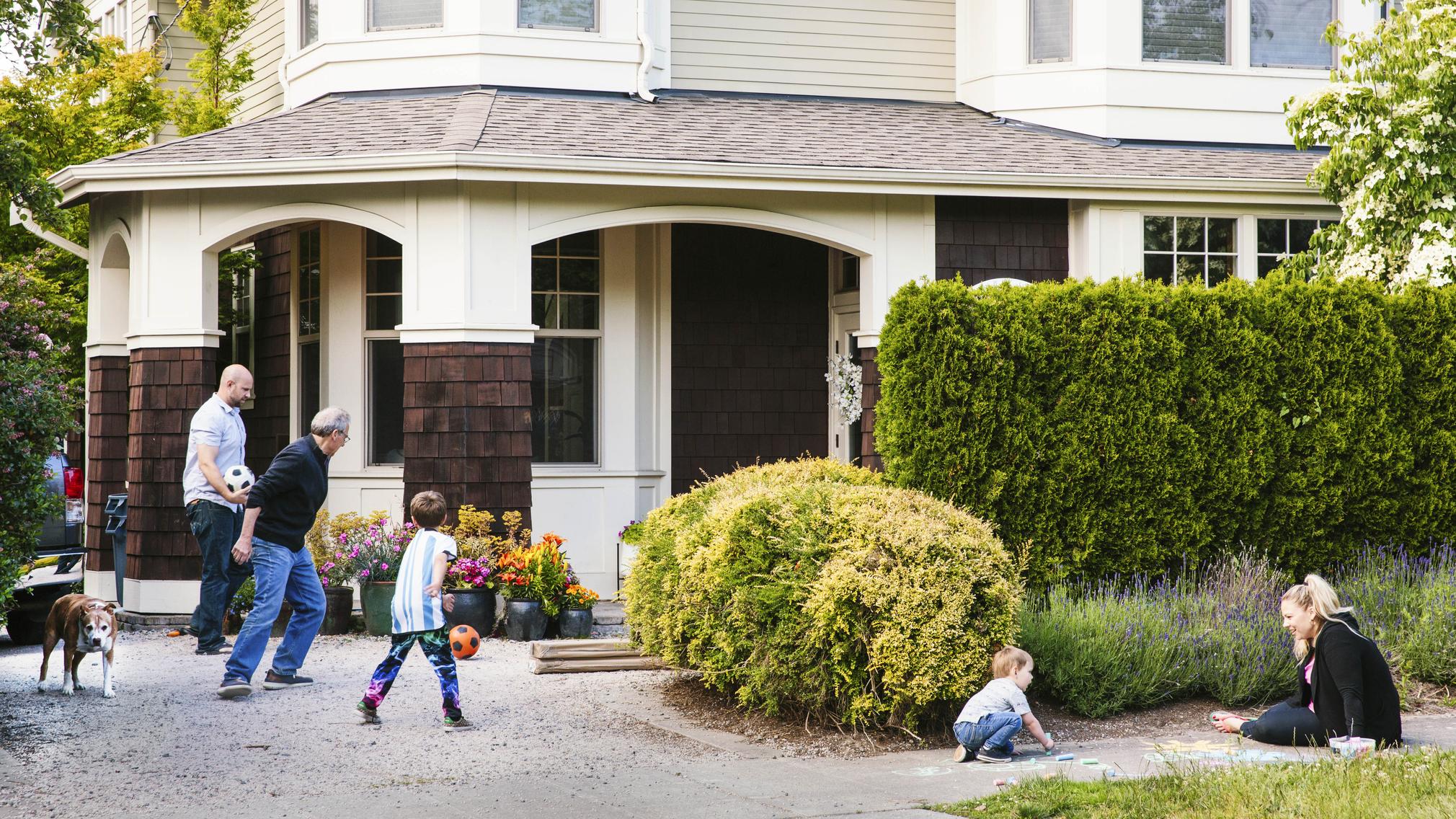 A multi-generational family of five, and their dog, play in the front yard of a beige Craftsman-style home. A grandfather, father and 7-year-old son play with a pair of soccer balls. Nearby, a mother sits on the sidewalk drawing with chalk along with her 2-year-old son.