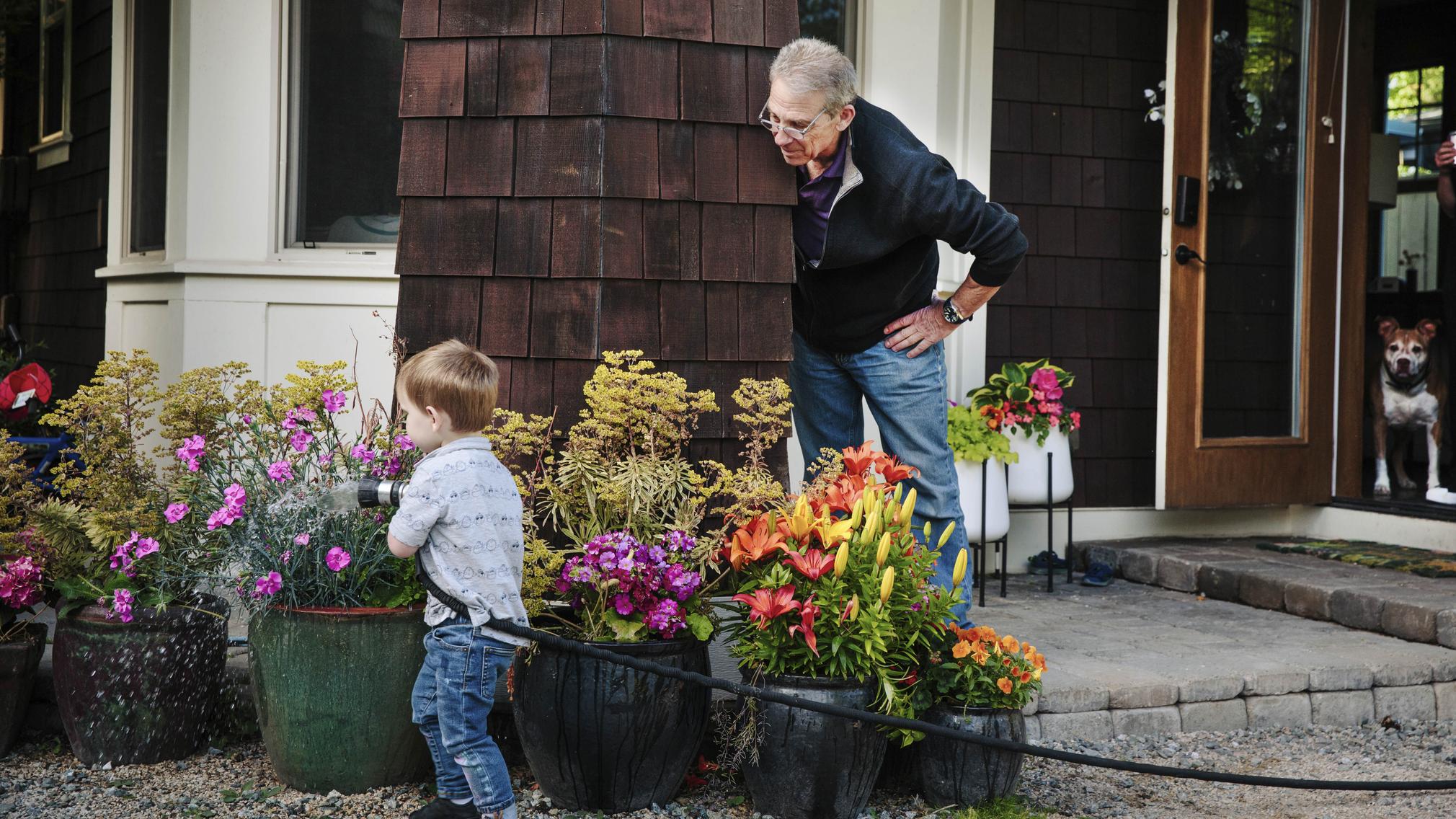 On the front porch of a Craftsman-style home, a man with grayed hair and glasses watches his 2-year-old grandson water various potted plants with a garden hose.