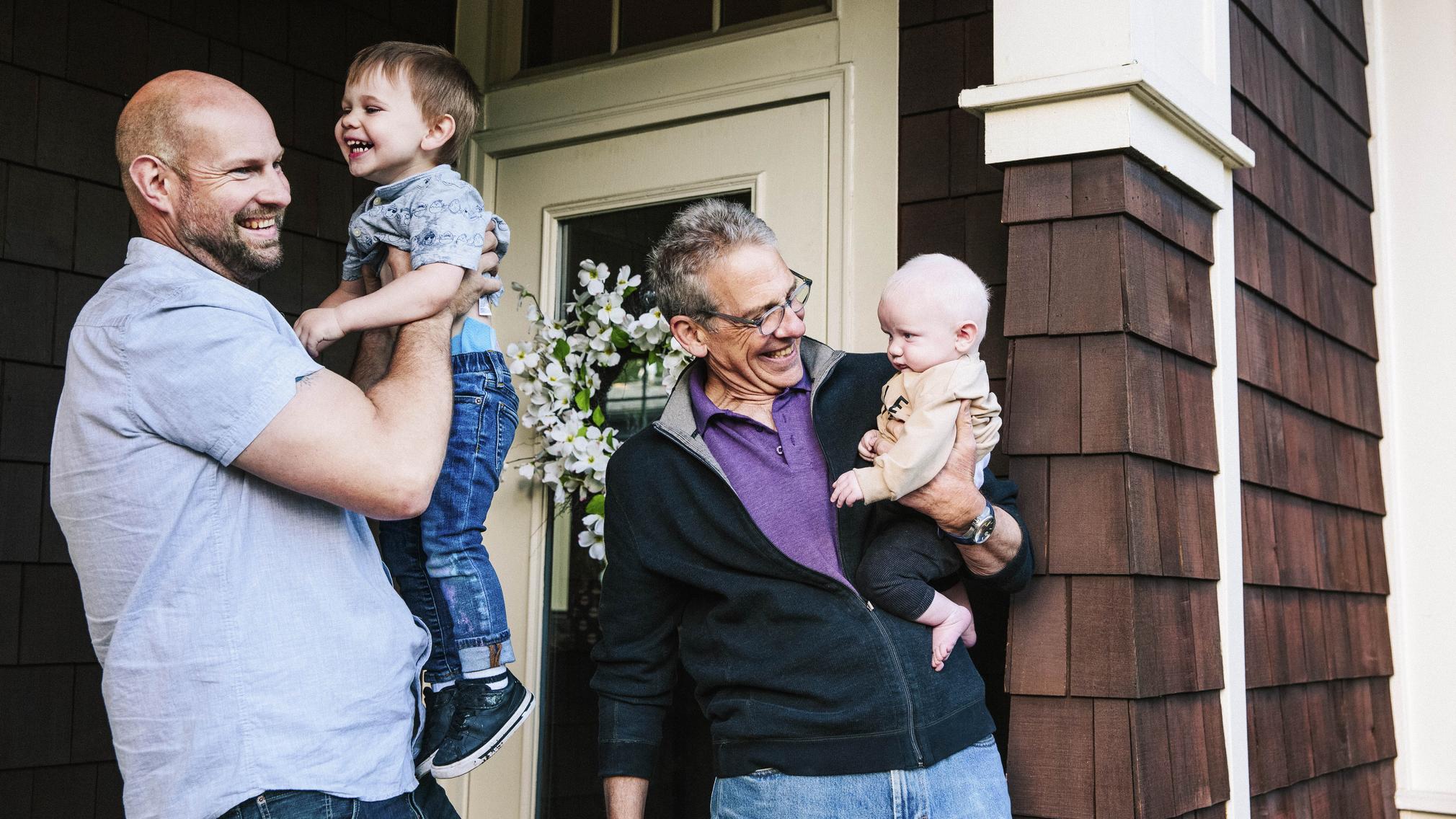 A father holds his 2-year-old son as the pair smile, and the boy's grandfather stands nearby holding and smiling at his infant brother.