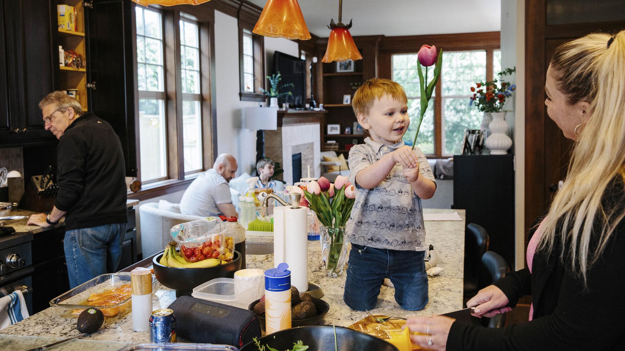 A 2-year-old boy kneels on a kitchen island, holding a tulip up to his mother, who smiles at him. There is a variety of food on the counter. In the background, the boy's grandson prepares dinner while a man and a 7-year-old boy sit in the living room.