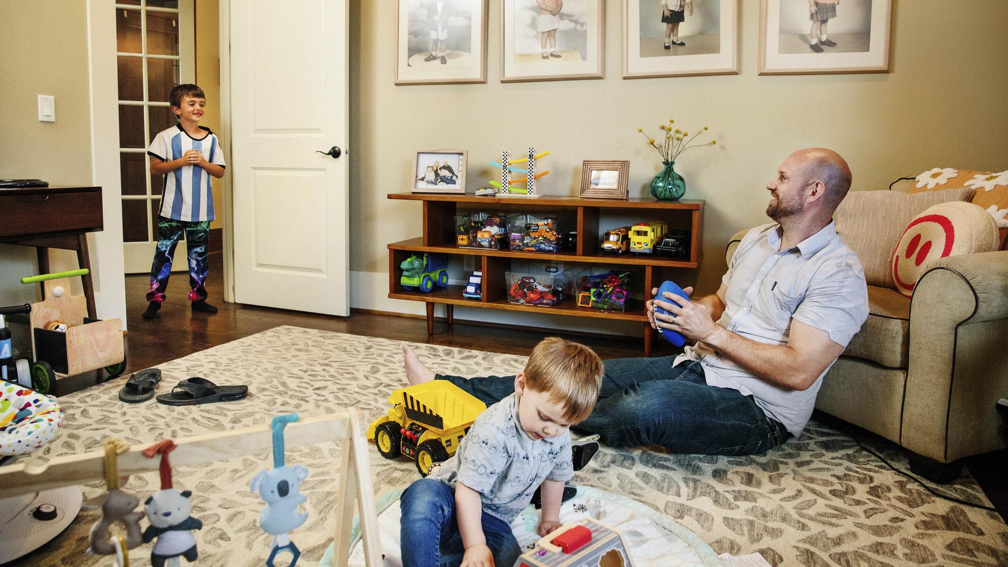 A middle-aged man sits on a rug in a beige living room, holding a foam football and smiling at his 7-year-old son. Meanwhile, his 2-year-old son plays with some toys nearby. There are toys throughout the room.