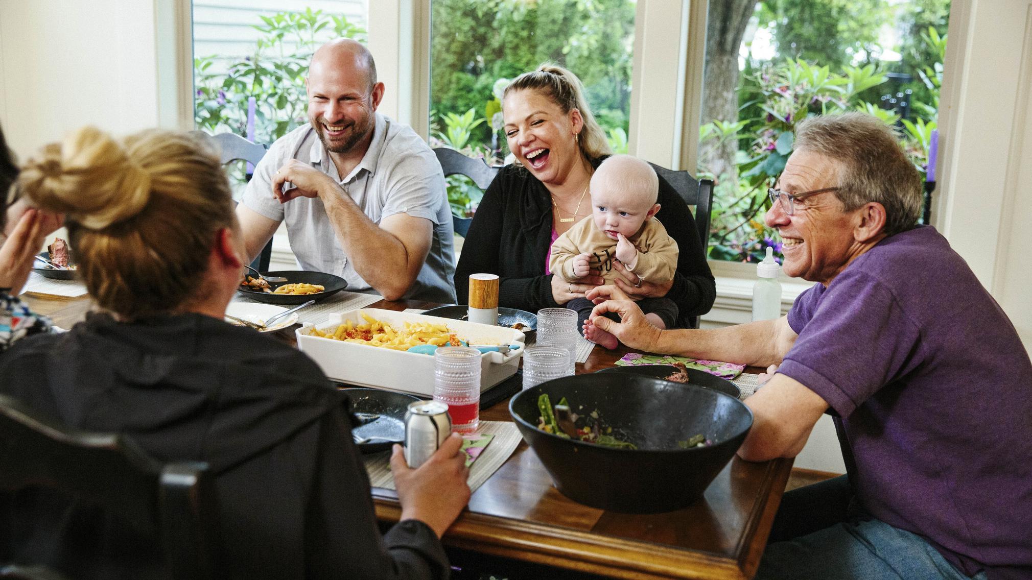 Four adults sit around a dining table, laughing mid-conversation. There are various large dishes for food to be shared from. A baby sits on his mother's lap and the child's grandfather holds the baby's foot.